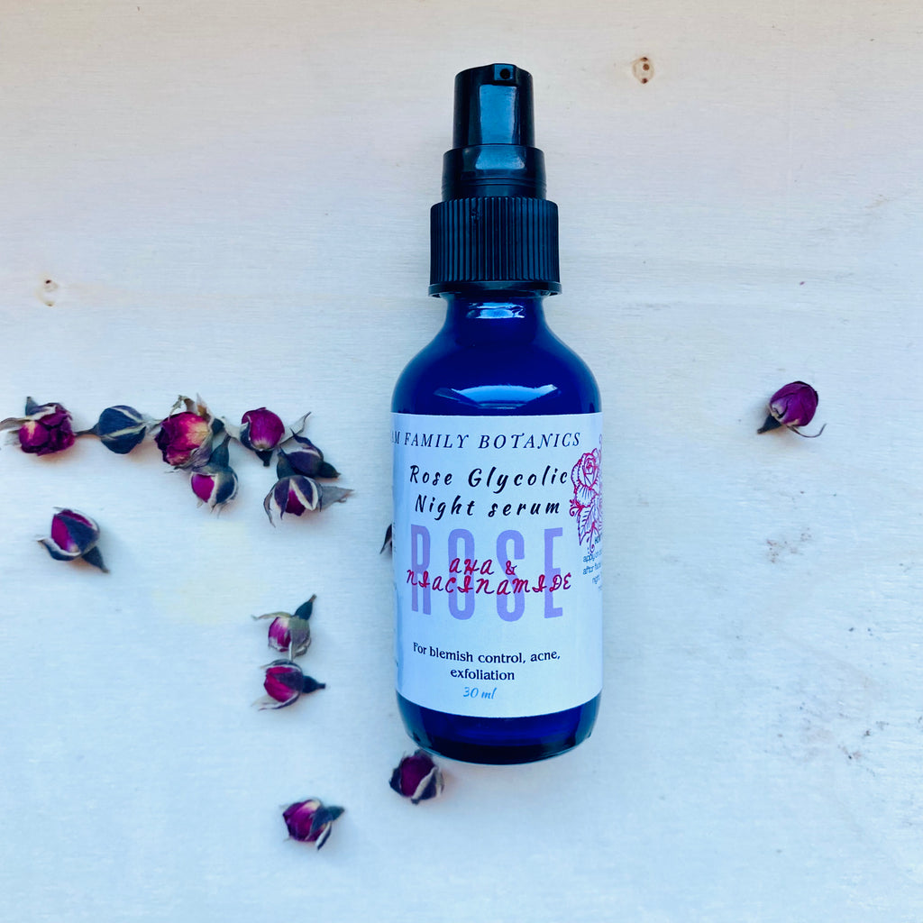 Blue glass bottle of Rose Glycolic Night Serum surrounded by small dried rosebuds on white background