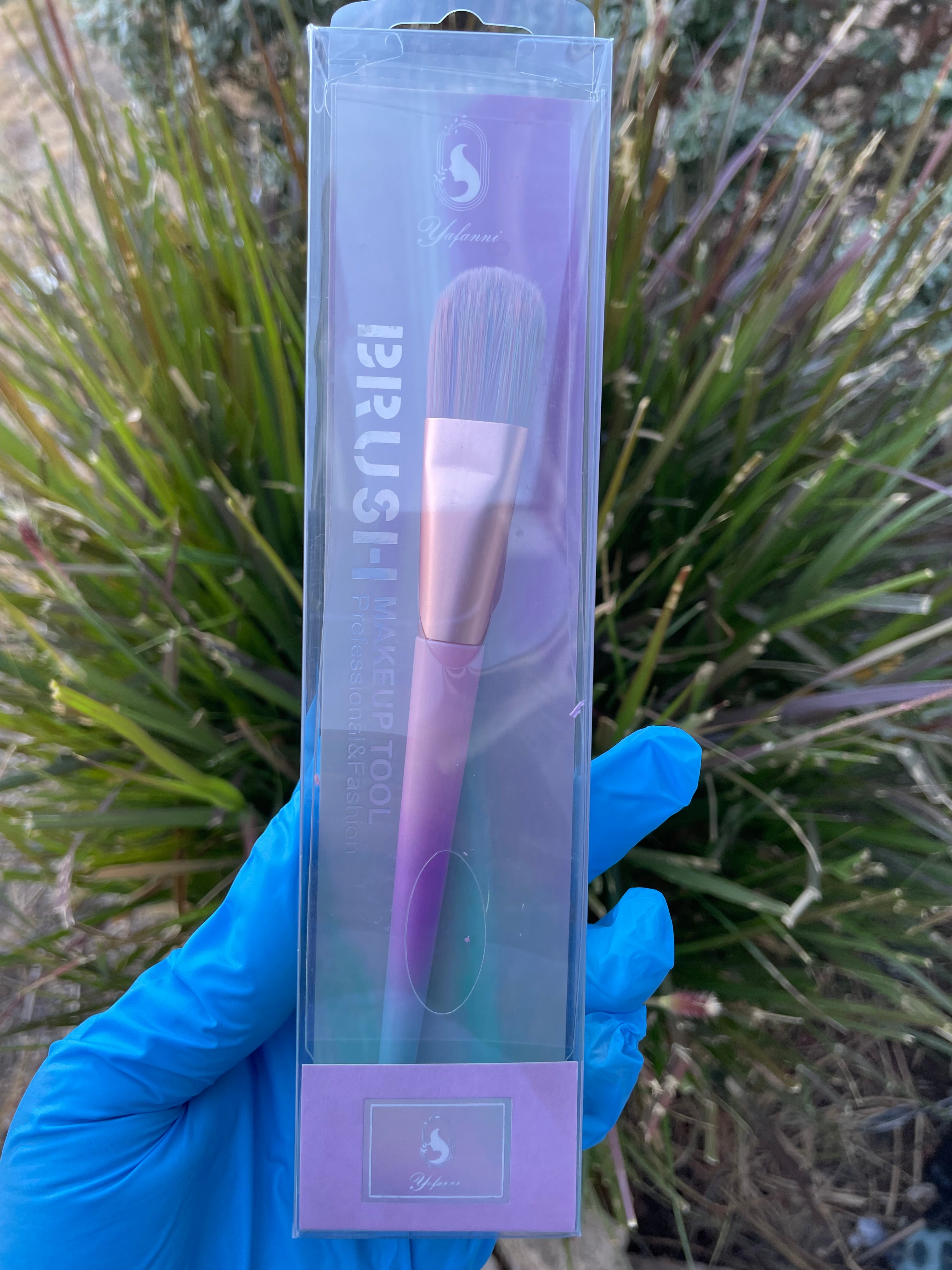 Unicorn Bronzer Brush with pastel handle and soft bristles in clear packaging held by gloved hand outdoors