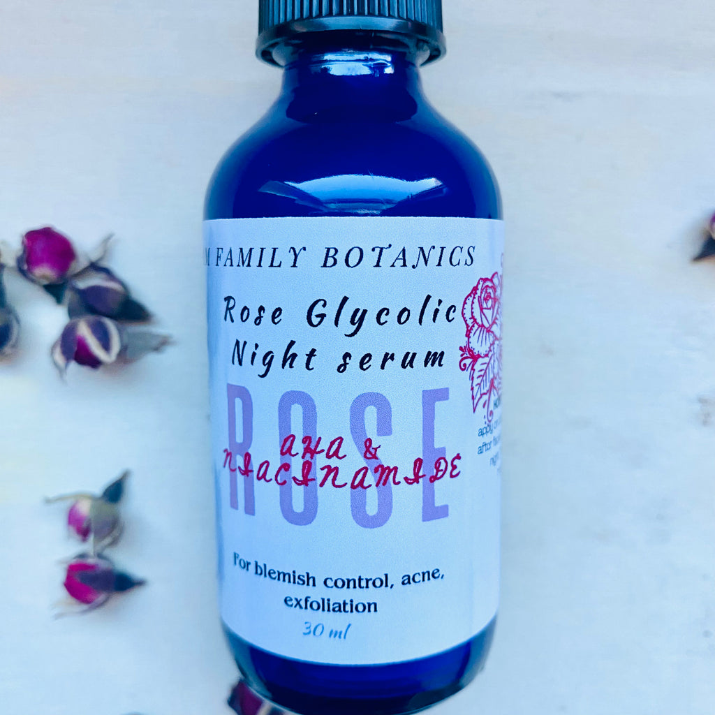 Blue glass bottle of Rose Glycolic Night Serum with rosebuds scattered on a white surface background