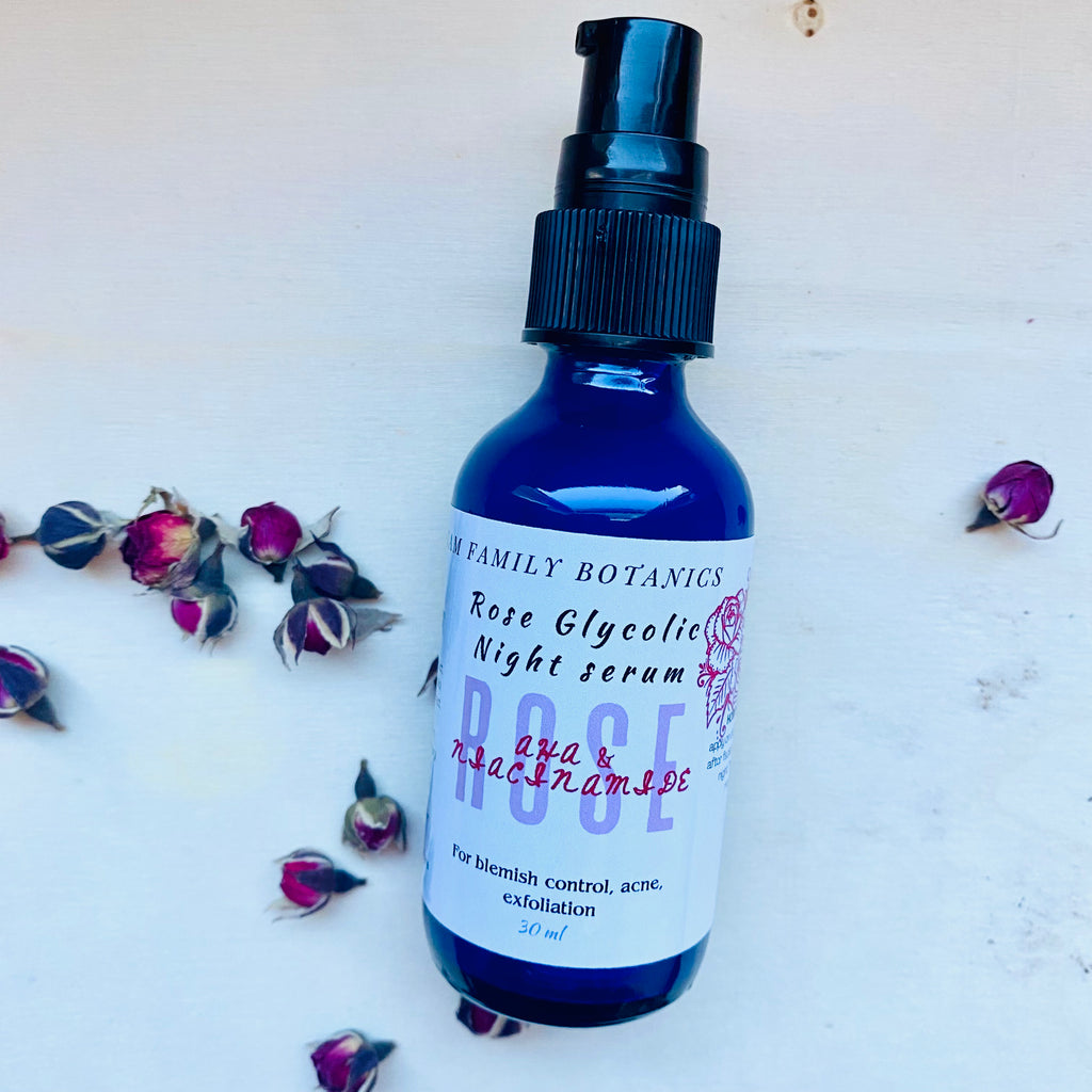 Blue bottle of Rose Glycolic Night Serum surrounded by small dried rosebuds on a white surface.