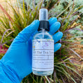 Hand in blue glove holding All The Tea Skin Toner Spray bottle outdoors with plants in background